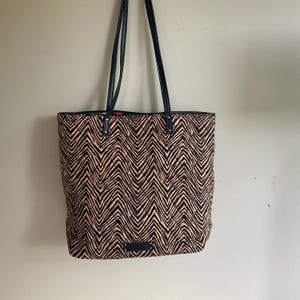 May include: A tote bag with a black and tan chevron pattern. The bag has black leather handles and trim. The bag is hanging against a neutral wall.