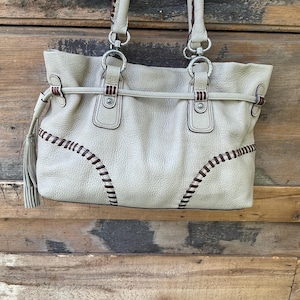 May include: Cream-colored leather tote bag with stitched detailing in dark brown. The bag features silver-tone hardware, double handles, and a tassel. The bag is set against a rustic wooden background.