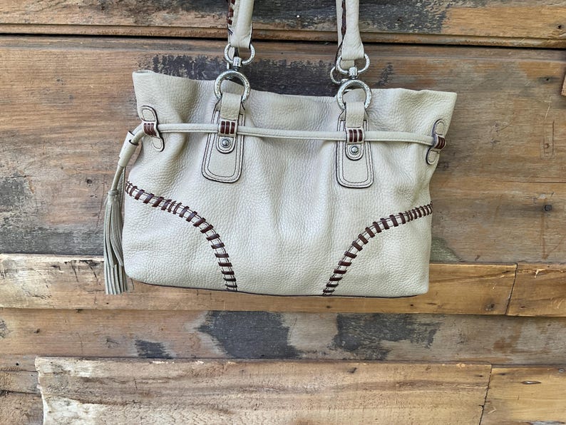 May include: Cream-colored leather tote bag with stitched detailing in dark brown. The bag features silver-tone hardware, double handles, and a tassel. The bag is set against a rustic wooden background.