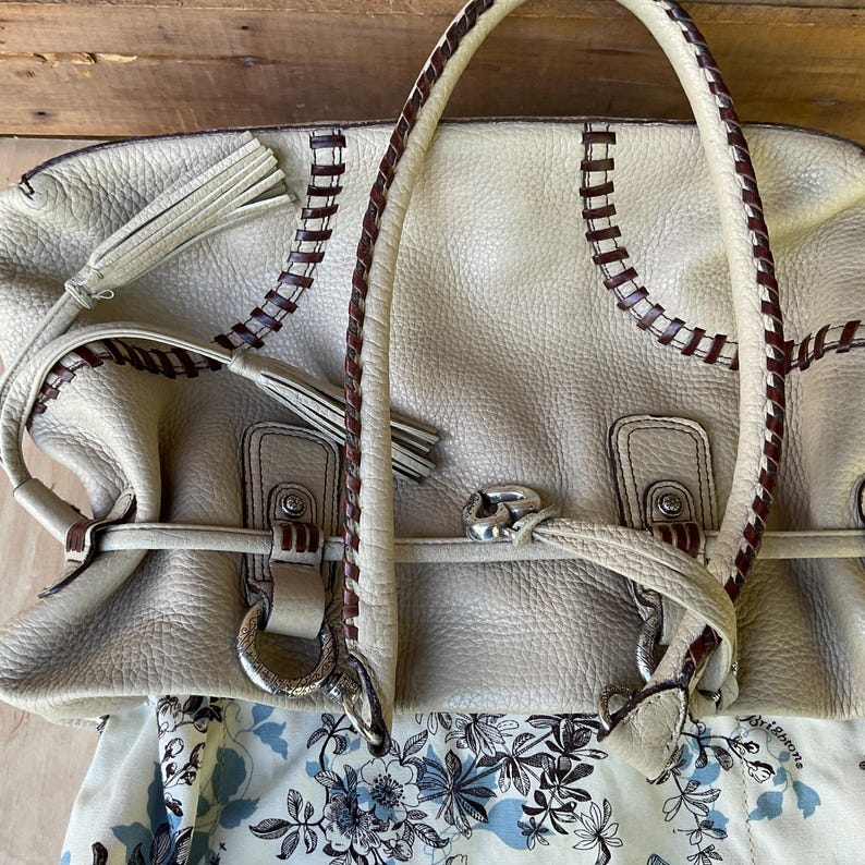 May include: Cream-colored leather handbag with brown stitching and braided handles. The bag features silver-toned hardware, tassels, and a floral patterned lining. A stylish and functional accessory.