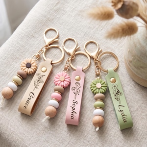 May include: Three personalised keychains featuring gold-tone metal rings and clasps. Each keychain has a leather-like strap engraved with a floral design and a name: Grace, Sophia, or Lydia. They include silicone flower beads and wooden beads.