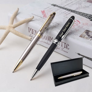 May include: Two silver and black pens with gold accents. The pens have the text "Your Text Here" engraved on them. One pen is in a black box.