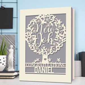 May include: A white card with a cut-out design of a tree with the words "New Job" inside the tree. The card has the words "Congratulations Daniel" at the bottom.