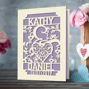May include: A white card with a purple background and a cut-out design of a heart, two birds, and the names Kathy and Daniel. The date 18/07/2019 is also cut out on the card.