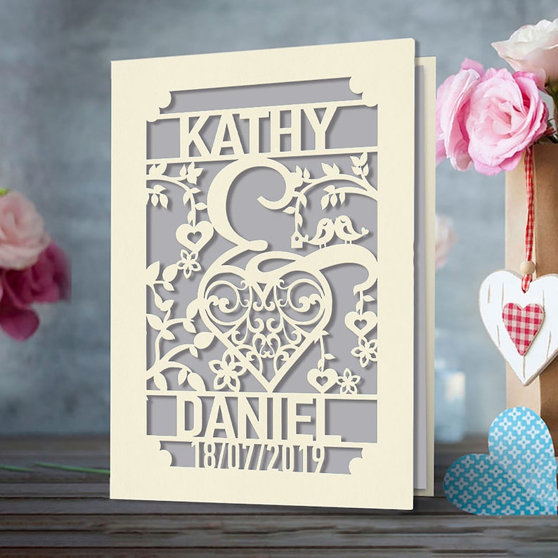 May include: A cream-colored card with a cut-out design featuring a heart, birds, and floral elements. The names "Kathy & Daniel" and the date "18/07/2019" are visible.