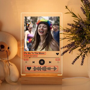 May include: A rectangular LED night light with a wooden base. The light displays a photo of a person and a music player interface. The text "Fly Me To The Moon" and "Frank Sinatra" are visible. The light is next to a stuffed bear.
