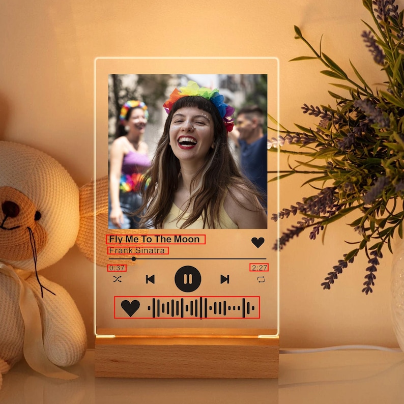 May include: A rectangular LED night light with a wooden base. The light displays a photo of a person and a music player interface. The text "Fly Me To The Moon" and "Frank Sinatra" are visible. The light is next to a stuffed bear.
