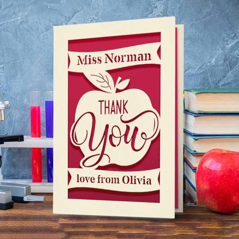 May include: A cream-colored card with a red apple cutout. The apple has the words "Thank You" inside. The card also has a banner that says "Miss Norman" and another banner that says "love from Olivia".