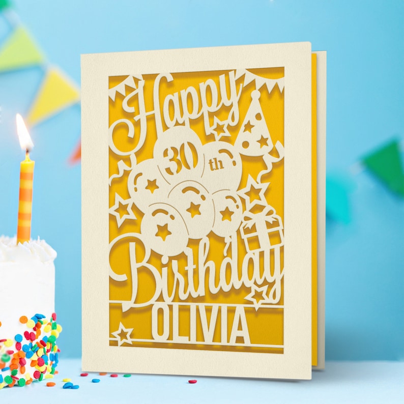 May include: A white card with a yellow background and a cut-out design of balloons, stars, a gift box, and a party hat. The text "Happy 30th Birthday Olivia" is cut out of the card.