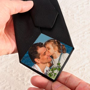 May include: Black tie featuring a photograph of a man and a child kissing. The tie is made of a textured black fabric. The photograph shows a man in a white shirt and a child in denim overalls. The background is a blue sky.