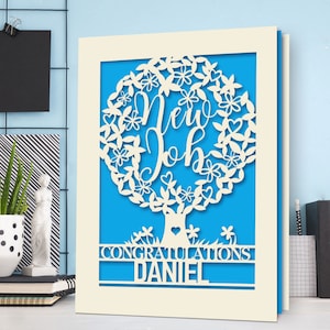May include: A white card with a blue background and a white paper cut-out design of a tree with the words "New Job" inside the tree. The card also has the words "Congratulations Daniel" at the bottom.
