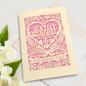 May include: A cream-coloured wedding invitation with a pink heart cutout. The heart features the names "Kathy & Daniel" and the date "19/06/2019". The invitation is decorated with a floral design and the words "Wedding Day" are printed on a banner.