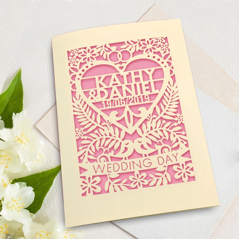 May include: A cream-coloured wedding invitation with a pink heart cutout. The heart features the names "Kathy & Daniel" and the date "19/06/2019". The invitation is decorated with a floral design and the words "Wedding Day" are printed on a banner.