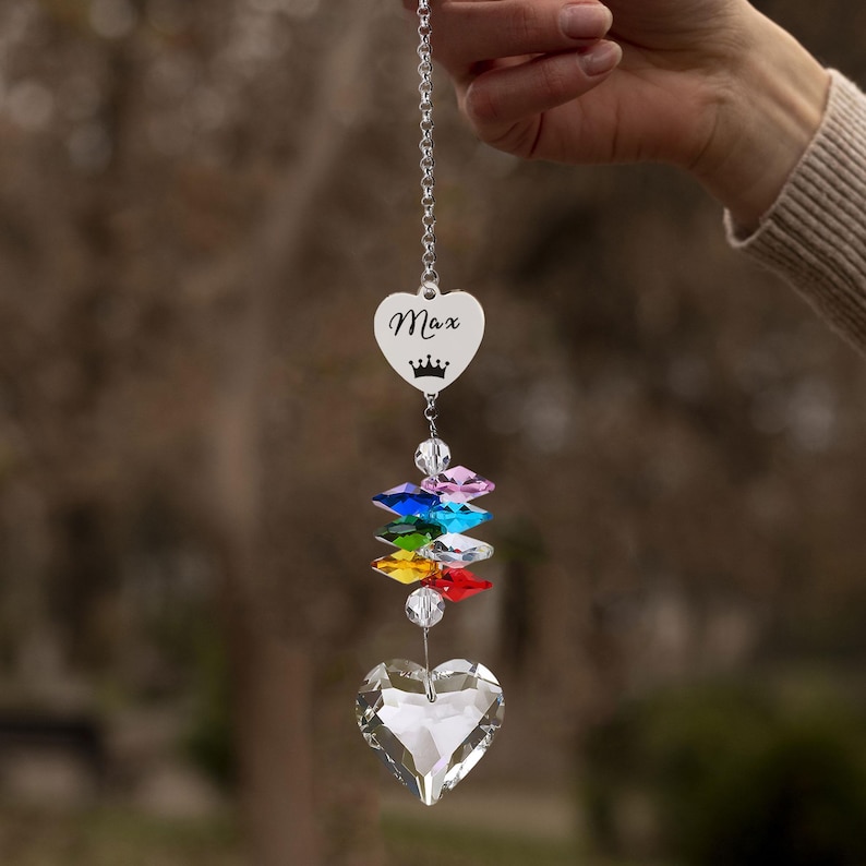 Puede incluir: Un atrapasoles de cristal en forma de coraz&oacute;n con el nombre "Max" y una corona grabados en un dije en forma de coraz&oacute;n. Debajo, un arco&iacute;ris de cristales de colores conduce a un gran cristal facetado en forma de coraz&oacute;n. El atrapasoles cuelga de una cadena plateada.