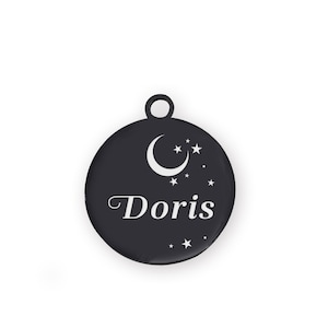 Puede incluir: Una placa redonda negra para mascotas con un dise&ntilde;o de luna creciente y estrellas. El nombre "Doris" est&aacute; escrito en blanco. La placa tiene un peque&ntilde;o lazo en la parte superior.