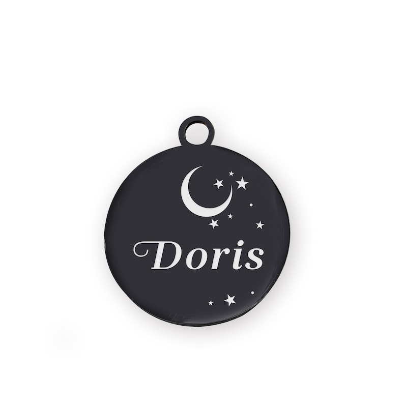 Puede incluir: Una placa redonda negra para mascotas con un dise&ntilde;o de luna creciente y estrellas. El nombre "Doris" est&aacute; escrito en blanco. La placa tiene un peque&ntilde;o lazo en la parte superior.