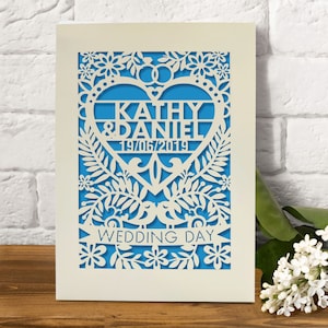 May include: A white card with a blue background and a heart-shaped cutout. The heart has the names "Kathy & Daniel" and the date "19/06/2019" inside. The card is decorated with a floral design and the words "Wedding Day" at the bottom.
