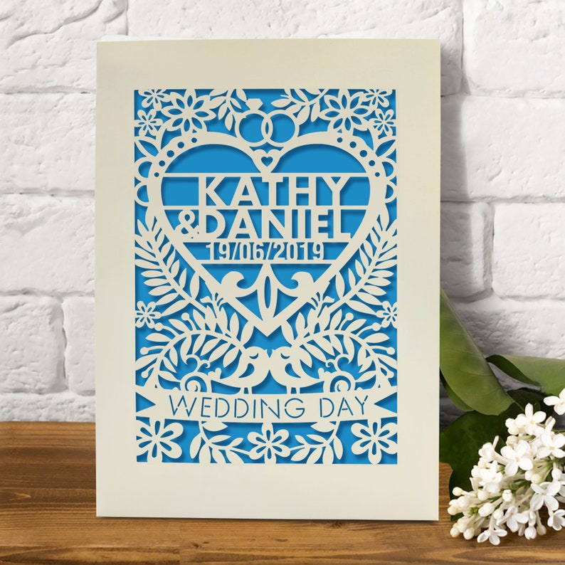 May include: A white card with a blue background and a heart-shaped cutout. The heart has the names "Kathy & Daniel" and the date "19/06/2019" inside. The card is decorated with a floral design and the words "Wedding Day" at the bottom.