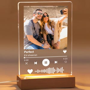 May include: A clear acrylic photo display with a wooden base. The display features a photo of a couple and a music player interface with the song title "Perfect" by Ed Sheeran. The base is rectangular and made of dark wood.