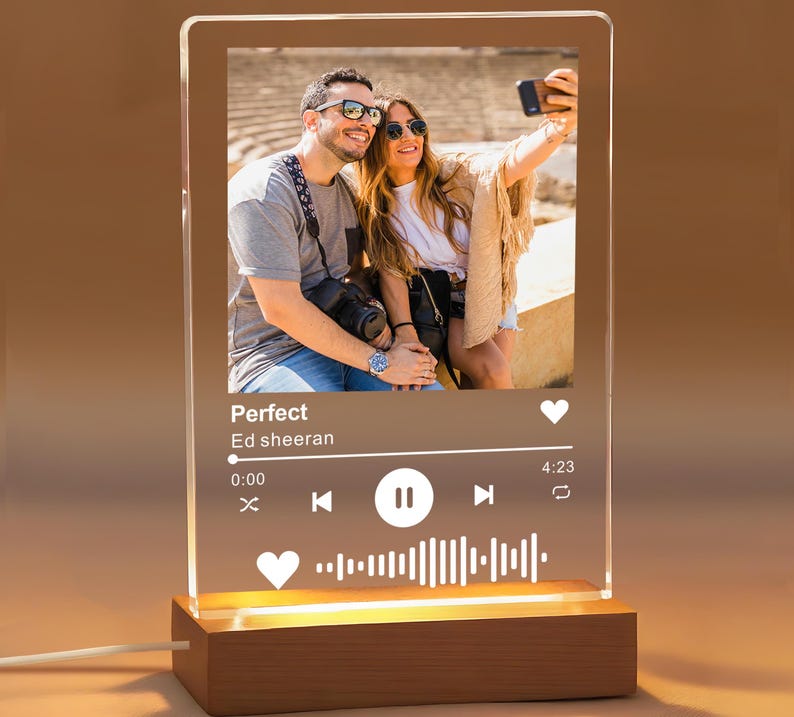 May include: A clear acrylic photo display with a wooden base. The display features a photo of a couple and a music player interface with the song title "Perfect" by Ed Sheeran. The base is rectangular and made of dark wood.