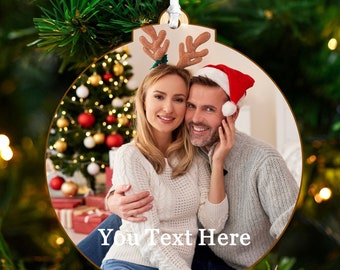 May include: A round wooden Christmas ornament with a photo of a couple in front of a Christmas tree. The couple is smiling and the man is wearing a Santa hat. The ornament has the text "You Text Here" printed on it.