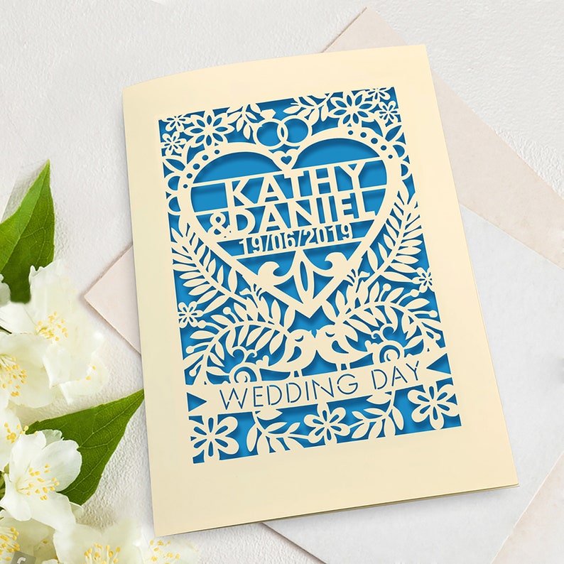 May include: A cream-coloured wedding invitation with a blue heart cutout. The heart features the names "Kathy & Daniel" and the date "19/06/2019". The invitation is decorated with a floral design and the words "Wedding Day" are printed on a banner.