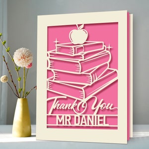 May include: A layered paper card with a pink interior and a cream-colored frame. The design features a stack of books topped with an apple, and the words "Thank You MR DANIEL" are cut out at the bottom. A vase of flowers is to the left.