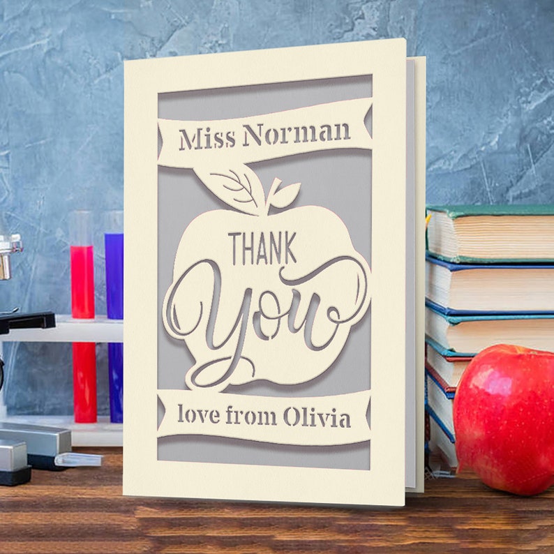 May include: A beige card with a cut-out design of a red apple with the words "Thank You" inside. The card also has a banner at the top that says "Miss Norman" and a banner at the bottom that says "love from Olivia".