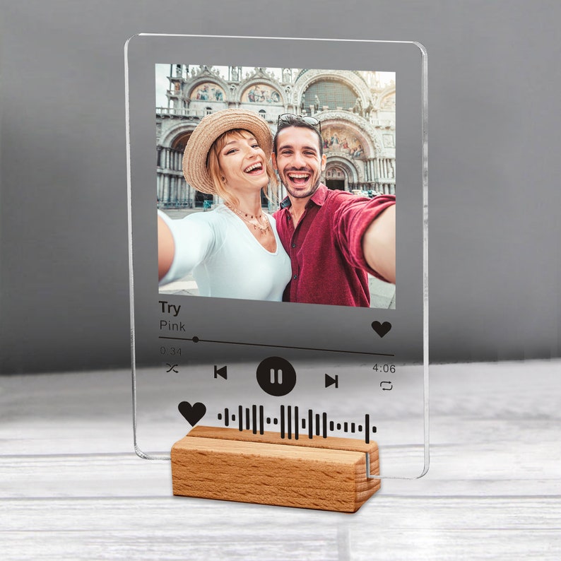 May include: Clear acrylic photo display with a wooden base. The display features a selfie of a smiling couple and a music player interface with the song title "Try" by Pink. The wooden base is rectangular and has a natural wood grain.