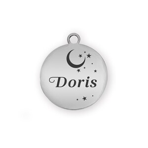 Puede incluir: Un charm redondo plateado con el nombre "Doris" en letra cursiva. El charm presenta un dise&ntilde;o de luna creciente y estrellas. Tiene un peque&ntilde;o lazo en la parte superior para sujetarlo.