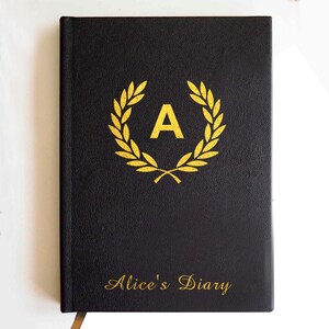 May include: A black hardcover journal featuring a gold-colored laurel wreath emblem surrounding the letter A in the center. The text Alice's Diary is printed in gold script at the bottom of the cover. The surface has a textured finish.
