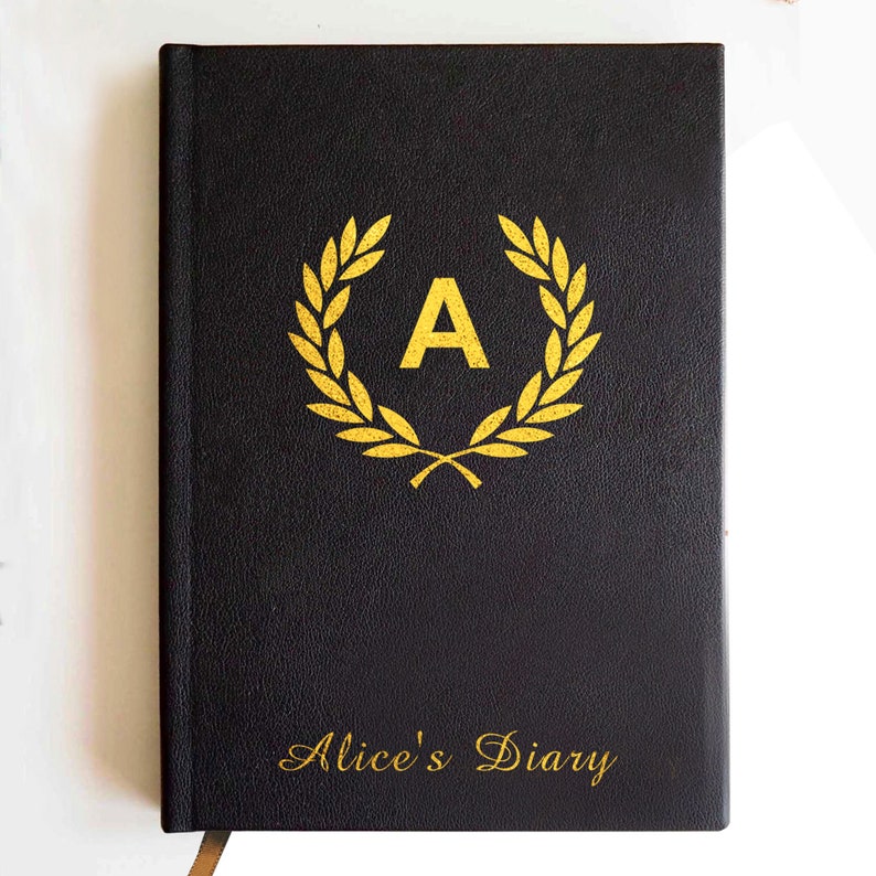 May include: A black hardcover journal featuring a gold-colored laurel wreath emblem surrounding the letter A in the center. The text Alice's Diary is printed in gold script at the bottom of the cover. The surface has a textured finish.