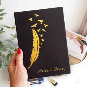May include: A black hardcover diary featuring a gold foil design of a single large feather with several small birds flying upward from the tip. The text Alice's Diary is printed in gold script at the bottom right corner of the front cover.