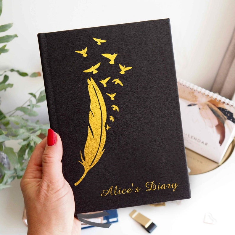 May include: A black hardcover diary featuring a gold foil design of a single large feather with several small birds flying upward from the tip. The text Alice's Diary is printed in gold script at the bottom right corner of the front cover.