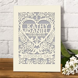 May include: A white card with a laser cut design of a heart with the words "Kathy & Daniel 19/06/2019" inside. The heart is surrounded by flowers and leaves. The words "Wedding Day" are written on a banner below the heart.