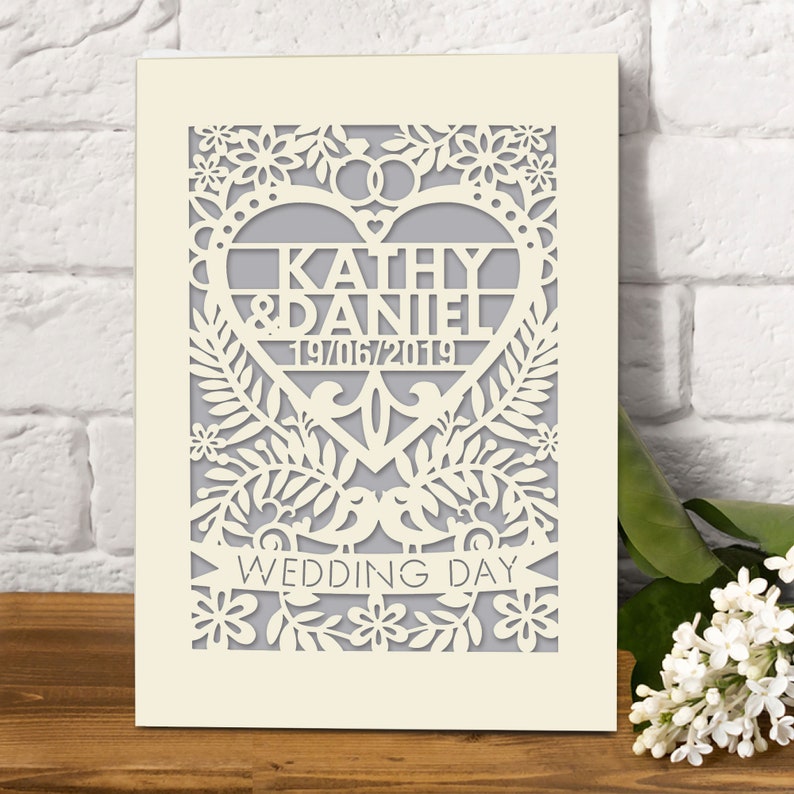 May include: A white card with a laser cut design of a heart with the words "Kathy & Daniel 19/06/2019" inside. The heart is surrounded by flowers and leaves. The words "Wedding Day" are written on a banner below the heart.
