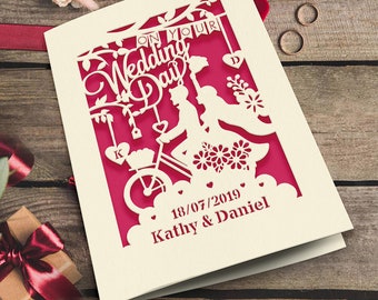 May include: A white wedding invitation card with a pink background. The card features a cut-out design of a couple riding a bicycle with flowers and hearts. The text on the card reads "On Your Wedding Day" and "18/07/2019 Kathy & Daniel".