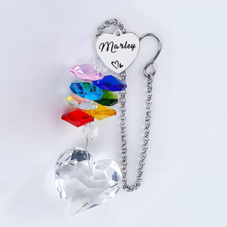 Puede incluir: Un atrapasoles de cristal en forma de coraz&oacute;n con un arco&iacute;ris de cristales de colores y una cadena plateada. El atrapasoles tiene una etiqueta en forma de coraz&oacute;n con el nombre "Marley" y un s&iacute;mbolo de coraz&oacute;n.