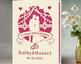 May include: A pink and white wedding card featuring a silhouette of a couple kissing under a string of lights. The card reads "Congratulations Kathy & Daniel 06.28.2022".