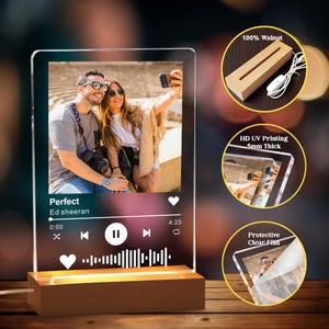 May include: A clear acrylic photo display with a wooden base, featuring a couple taking a selfie. The display includes a music player interface with the song title "Perfect" by Ed Sheeran. The base is made of walnut and has a warm light.