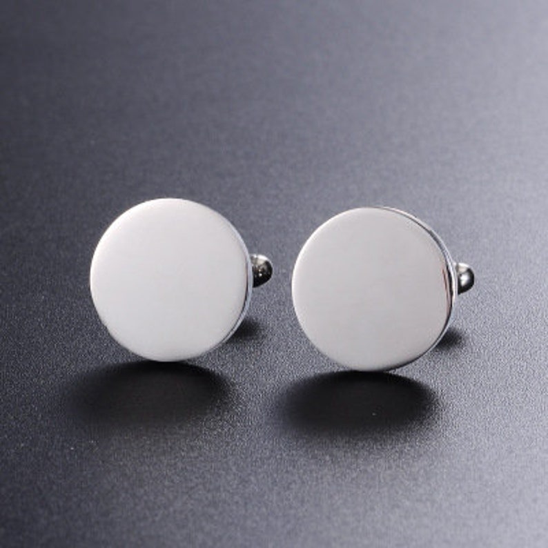 May include: A pair of silver cufflinks with a round, smooth, blank surface.