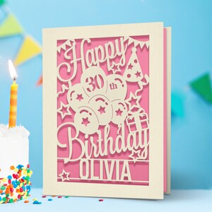 May include: A white card with a pink background and a cut-out design of balloons, stars, and a gift box. The text "Happy 30th Birthday Olivia" is cut out of the card.