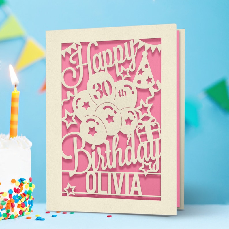 May include: A white card with a pink background and a cut-out design of balloons, stars, and a gift box. The text "Happy 30th Birthday Olivia" is cut out of the card.