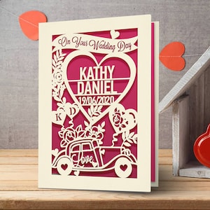 May include: A white card with a pink background and a cut-out design of a car with flowers and a heart with the text "Kathy Daniel 19/06/2020" and the text "On Your Wedding Day" above the heart.