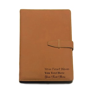 May include: A brown leather journal with a strap closure. The journal has the text "You Text Here" engraved on the front.