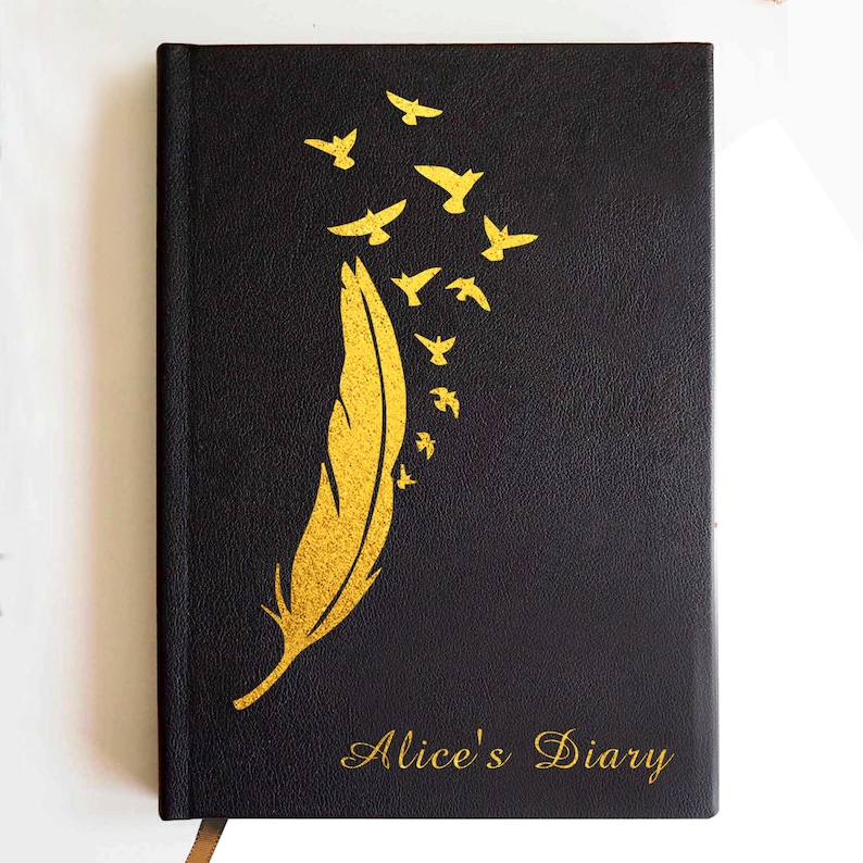 May include: A black hardcover journal featuring a gold foil design of a single large feather transforming into a flock of small birds flying upward. The text Alice's Diary is printed in gold script at the bottom right corner of the front cover.