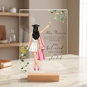 May include: A clear acrylic plaque on a wooden base featuring a printed illustration of a graduate in a white dress and pink gown holding a diploma and sunflowers. Text reads: Abby Thomson, University of Nottingham, PG Cert Medical Education, Class of 2026, She believed she could so she did.