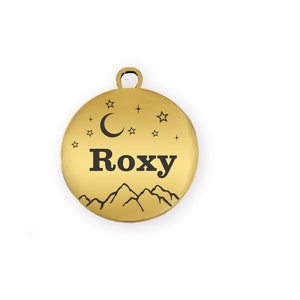 Puede incluir: Una placa redonda dorada para mascotas con el nombre "Roxy" en letras negras en negrita. La placa presenta un dise&ntilde;o de luna creciente, estrellas y cordillera. Tiene un peque&ntilde;o lazo en la parte superior para sujetarla.