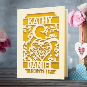 May include: A white card with a yellow background and a cut-out design of a heart with vines, birds, and flowers. The names "Kathy" and "Daniel" are at the top and the date "18/07/2019" is at the bottom.