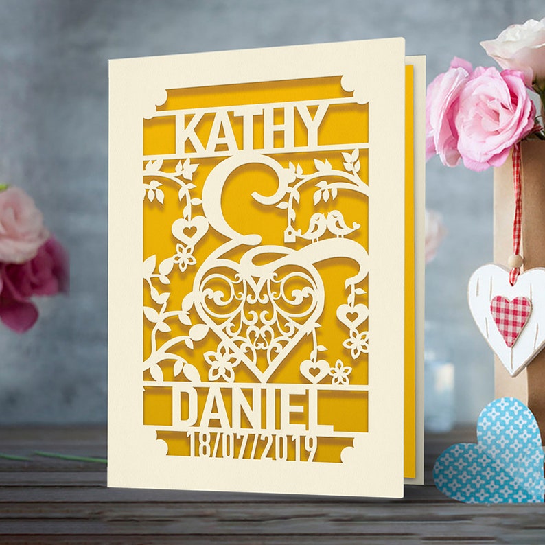 May include: A white card with a yellow background and a cut-out design of a heart with vines, birds, and flowers. The names "Kathy" and "Daniel" are at the top and the date "18/07/2019" is at the bottom.